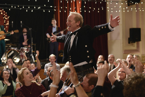 A man in a full suit and waistcoast stands above a crowd at a wedding, in a still from Sunshine on Leith.