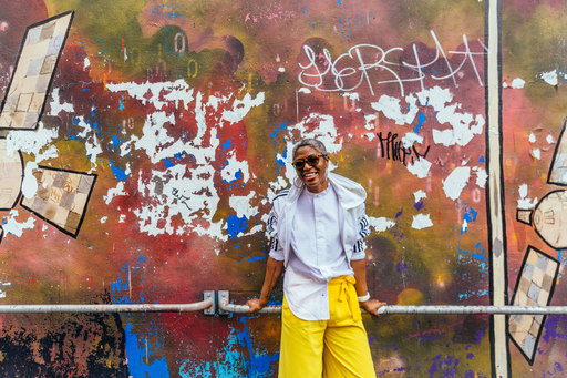 DJ Paulette, standing in front of a wall with patches of paint and graffiti.