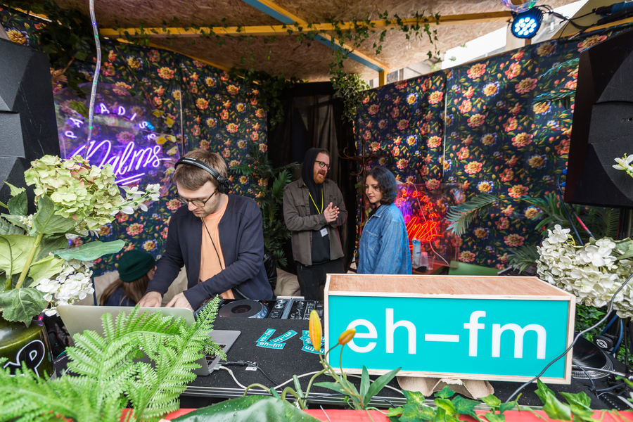 A DJ performs from a booth decorated with plants. An EHFM lightbox is next to the DJ equipment; a man and a woman are having a conversation at the back of the booth.