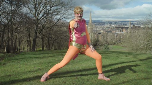 A man in a pink and orange superhero outfit stands on a hill overlooking a city skyline; he is pointing directly at the viewer.