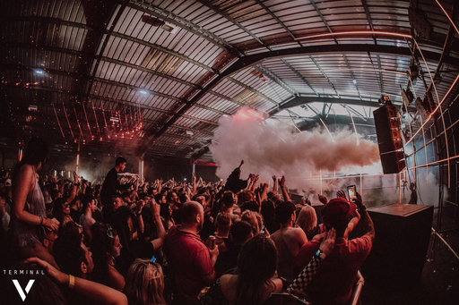 Crowds gather in front of an indoor stage, surrounded by red lights and smoke