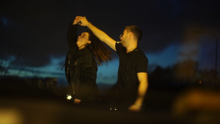 Two figures dressed in black, dancing. The male figure on the right is smoking a cigarette.