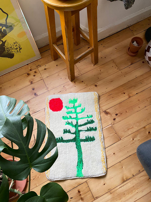 A pale rug decorated with a bright green tree and a red sun sits on a wooden floor. A cheeseplant is in the foreground, with a stool and various art materials placed at the edges of the frame.