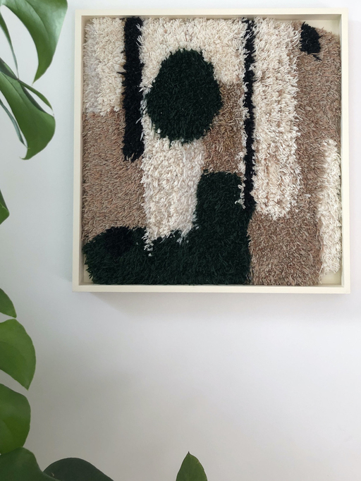 A rug hanging on a wall. The rug is brown with interlocking black and white figurative shapes emerging from the top and bottom. A plant is visible to the left of the rug.