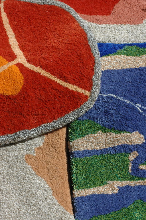 Three overlapping rugs. In the top left, a red rug with a grey border and white and yellow detailing. On the right, a blue and green rug with grey striations across it. In the bottom left, a pink and grey patterned rug.