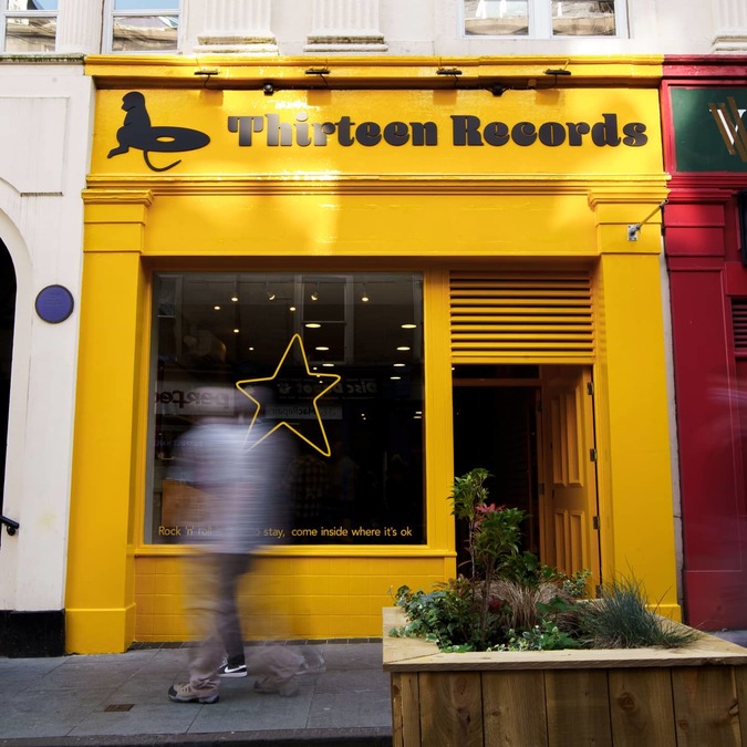 The exterior of Thirteen Records; a yellow painted single-story shopfront, with a large window decorated with a yellow star. A member of the public walks past in the foreground.