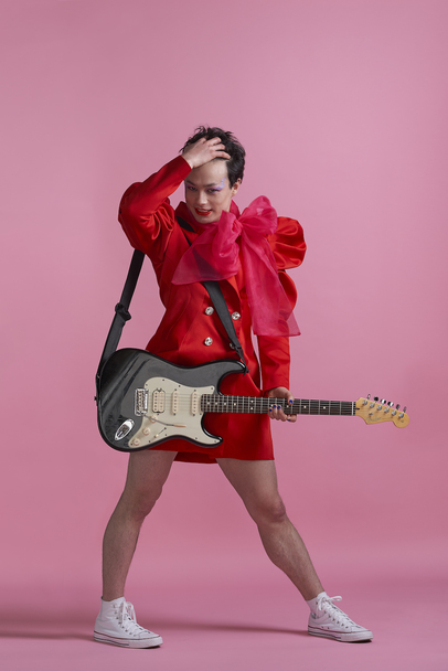 Portrait photo of Sean Shibe. Sean stands against a pink backdrop holding a black electric guitar, wearing a mid-length red coat with a large pink bow on the left shoulder. 