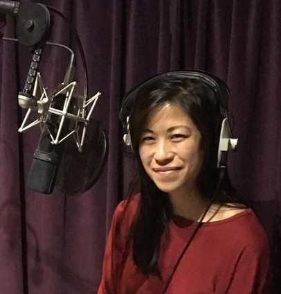 Anna Cheung in what appears to be a recording studio. Anna wears headphones, with a microphone set up in front of her and purple velvet curtains behind.