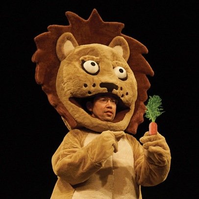 Mamoru Iriguchi, dressed in an oversized lion costume. His face is visible within the lion's mouth, and he holds a prop carrot.