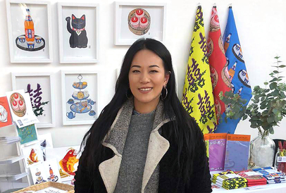 Sarah Kwan stands in front of an array of framed works and assorted stationery.