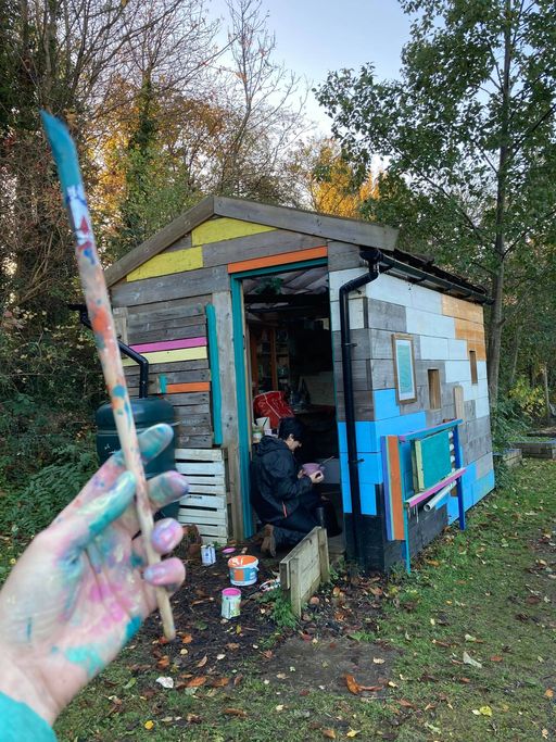 A multi-coloured shed in a wooded area; photographer is holding a paint-covered paintbrush in the foreground, while another person retrieves objects from inside the shed.