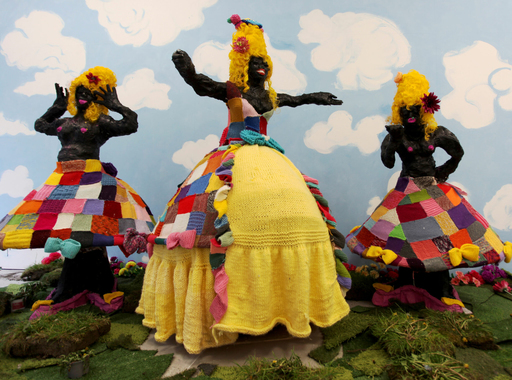 Installation view of work by Josie KO - three large black papier-mache models are posed in multi-coloured patchwork dresses, against a background of blue sky and white clouds