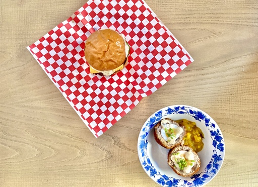 Photo of food from King of Feasts. A cheeseburger sits on a red and white-checked wrapper, and a scotch egg is in a white porcelain bowl with a blue floral motif around its edge.