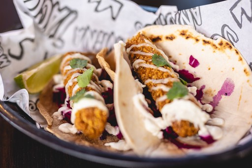 Two tacos in a paper lined basket, filled with fried vegetables and topped with herbs and red onion.