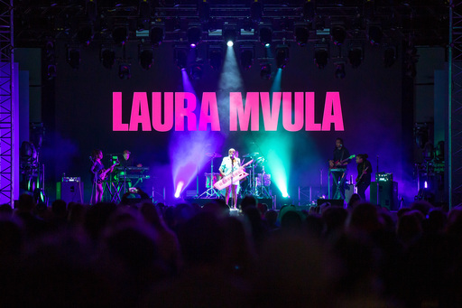 Laura Mvula performs at Edinburgh International Festival. A crowd stands in front of the stage; Mvula stands in the centre holding a keytar with various musicians around her. The words 'Laura Mvula' are projected on the screen behind her.