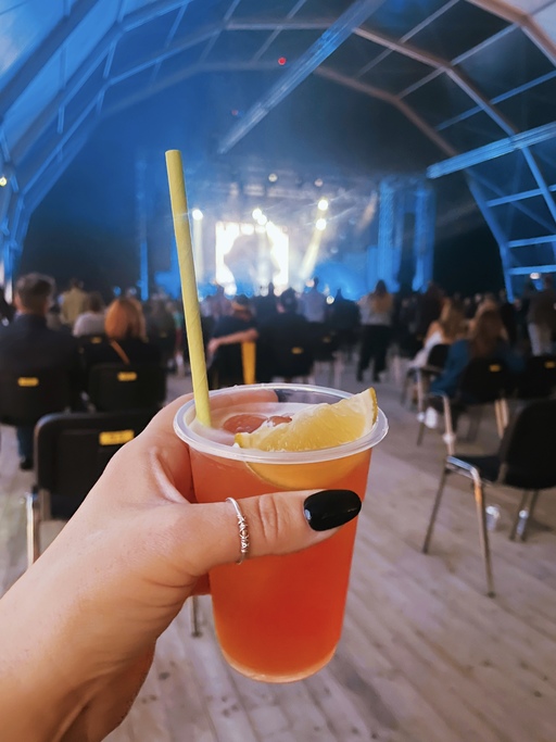 A hand holds an orange drink in front of a concert stage.