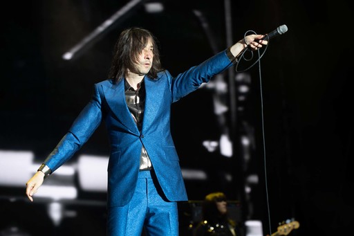 Bobby Gillespie of Primal Scream holds his microphone towards the audience while on stage at TRNSMT festival. He is dressed in a bright blue suit.