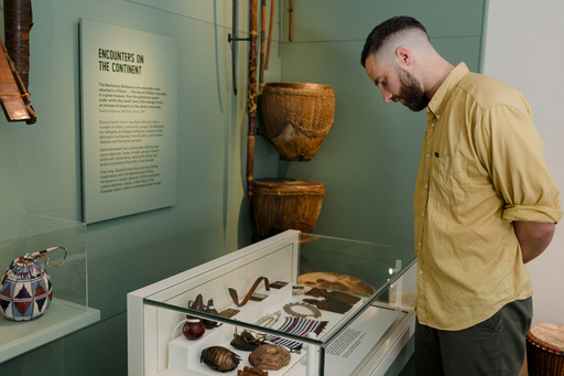 A man in a yellow shirt stands over a museum cabinet filled with artefacts and objects. Two large drums hang from the wall by the cabinet. Text on the wall panel is ineligible, but title reads 'Encounters on the Continent'