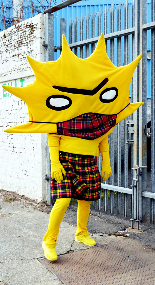 Kingsley, the mascot of Partick Thistle; a yellow figure with a head shaped like a sunburst. He is wearing a kilt and a tartan facemask, standing in front of a locked gate.