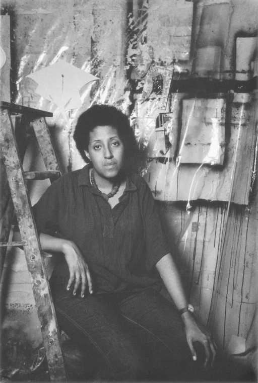 A black and white photograph of Howardena Pindell. She wears a dark blouse and jeans, and is leaning on a paint-splattered ladder.