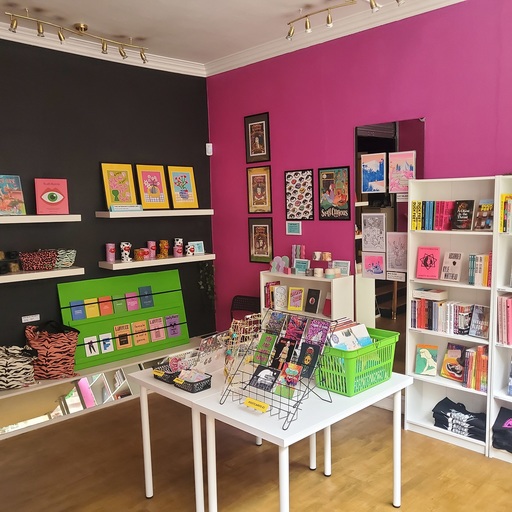 Interior view of Blunt Knife Co; a central table is covered with objects and books, with pink walls behind and shelves filled with books, clothing and various objects. 