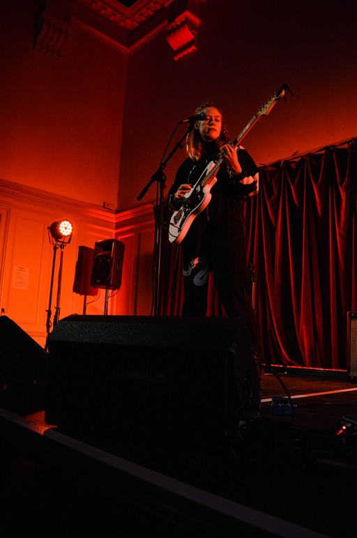 Anna B Savage stands on an indoor stage surrounded by red lights as she performs with a guitar