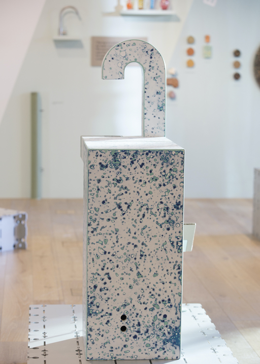 A drinking fountain with a tall, thin rectangular base and long curved spout, made from a white material with blue mottled patches.