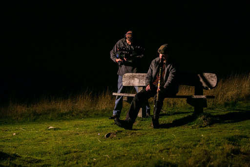 A camera operator films a man playing a didgeridoo on a park bench.