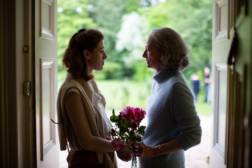 A young woman stands opposite her mother in a doorway, holding a bunch of flowers, in a still from The Souvenir Part 2
