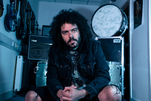 Matthew Hickman aka brownbear sitting in front of a collection of guitars and amplifiers.