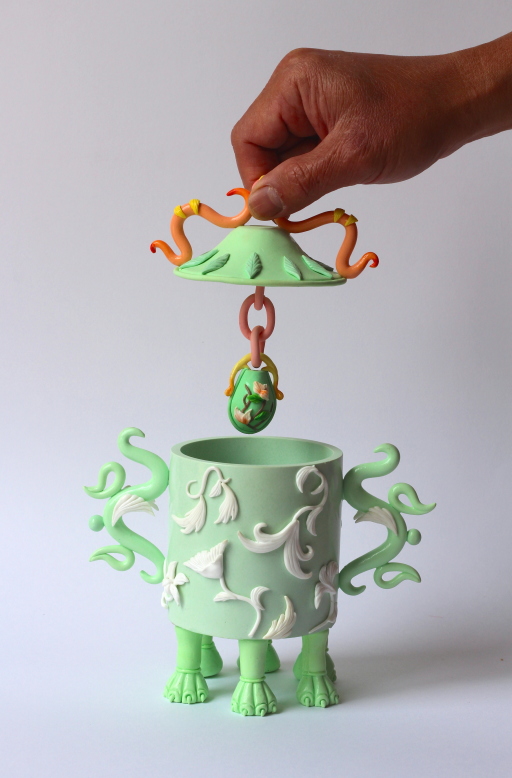 A small green pot with six feet, and ornate arms on either side. There is an egg-shaped object hanging from the pot's lid, which is being held aloft.