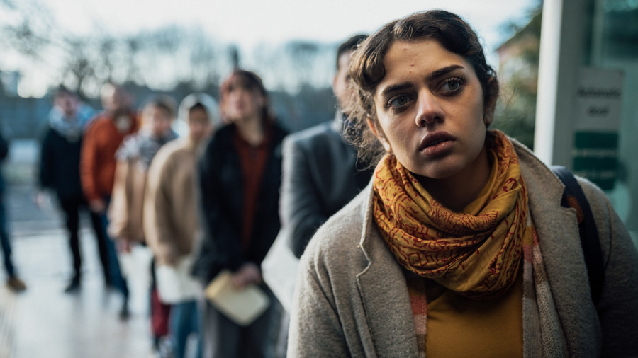 A woman in a grey coat and gold scarf stands at the front of a queue of people.