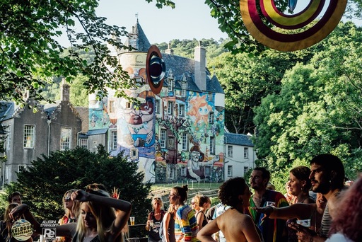 A crowd of people stand in front of Kelburn Castle.  The building is surrounded by trees and the walls are painted with colorful murals