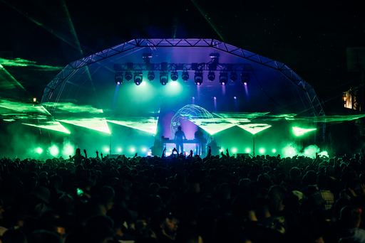 An electronic artist performs for a large crowd under blue and green lights