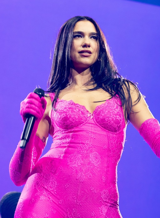 Dua Lipa stands on stage in a pink bodysuit, holding a microphone.