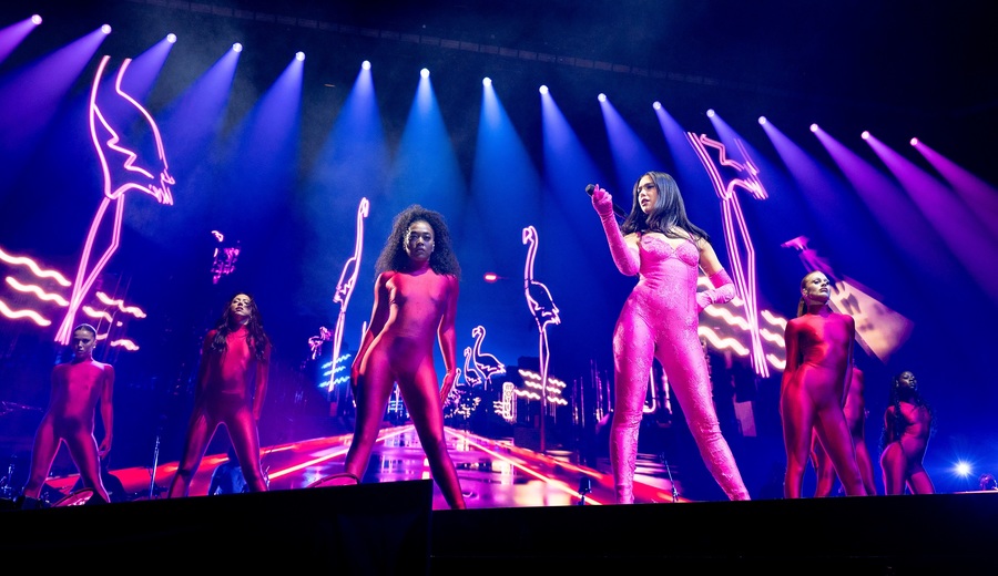 Dua Lipa stands on stage in a pink bodysuit, holding a microphone. Dancers in red bodysuits stand behind her, while pink neon lights are visible behind the group.