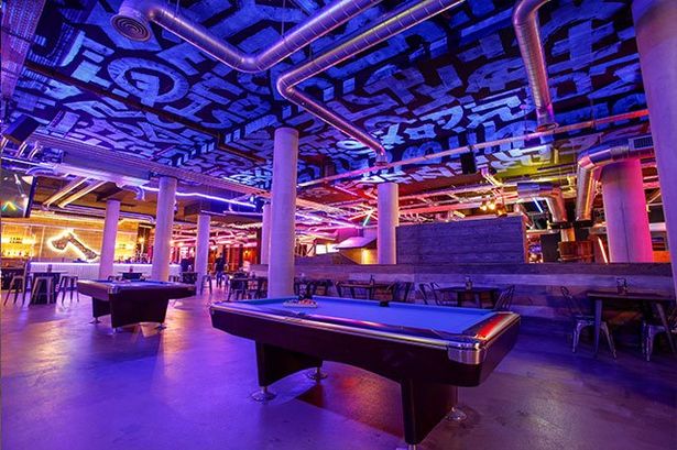 Pool tables are spaced out under blue and magenta lighting. In the distance there are bar stools and tables, as well as an axe-throwing activity space. The ceiling is decorated in blue glyphs.