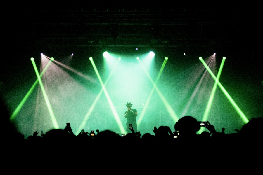 Denzel Curry on stage, with green lights shining around him; he is framed by silhouetted members of the audience holding up their camera phones.