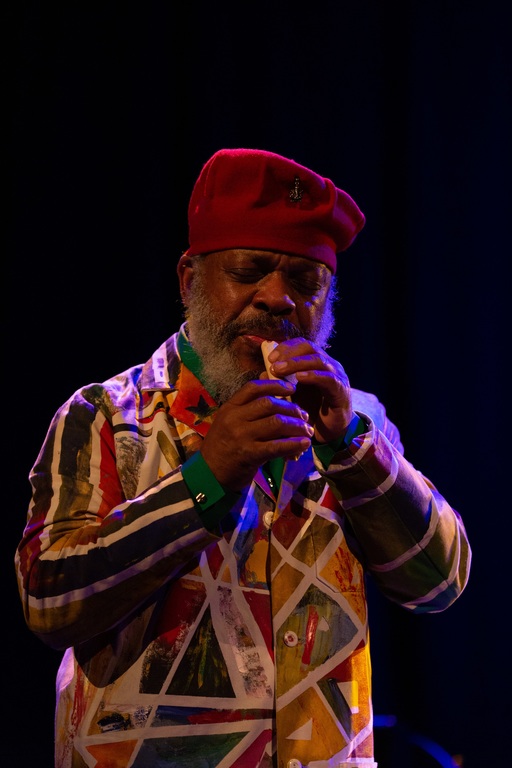 Douglas R Ewart, wearing a red felt hat and a suit covered in multicoloured geometric shapes, plays a small whistle.