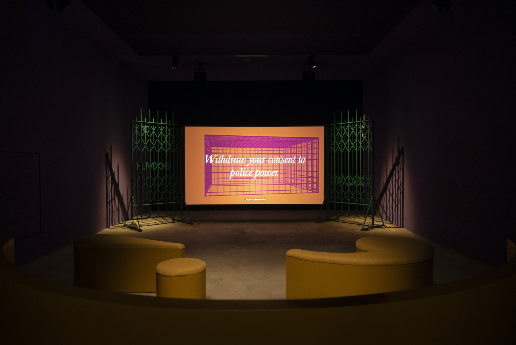 A video screen framed by two green metal gates, in front of a selection of yellow chairs. Screen shows a pink grid against an orange background, overlaid text reads 'Withdraw your consent to police power.'