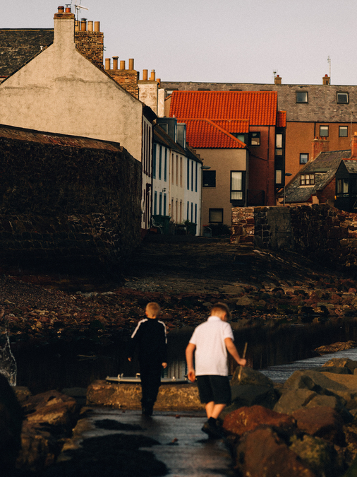 Two children cross a stream, beyond which is the brick buildings of Dunbar