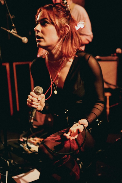 Vicky Kavanagh of Bikini Body on stage, holding a microphone and looking off-screen. A band member with a guitar stands behind her.