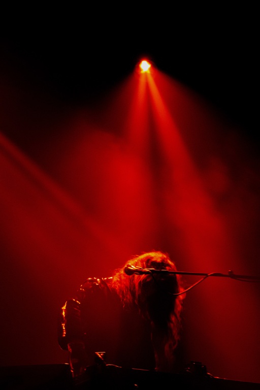 A member of Beach House on stage at Glasgow Barrowlands. Hair covers their face, and they are obscured by the red stage lights.