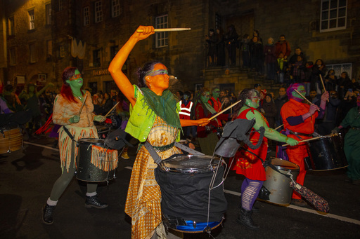 A group of drummers wearing green paint march past a crowd on an Edinburgh street.