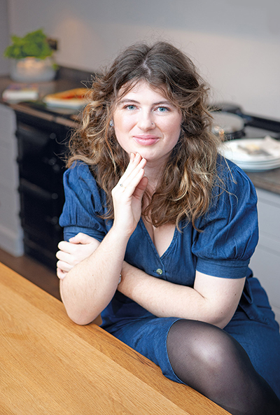 Ella Risbridger sits at a kitchen table, wearing a blue dress, with dishes visible in the background.