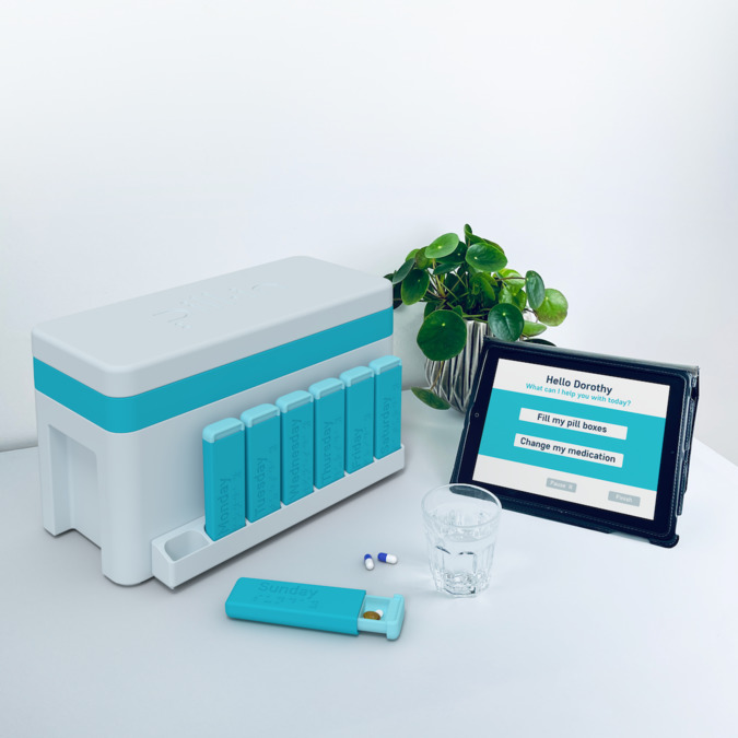 A white and teal box, with a video screen showing user options to fill pill boxes or change medication. A green plant sits on the table behind the device.