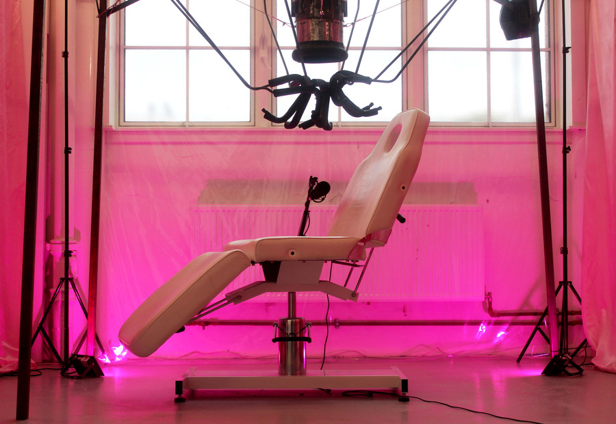 A pastel pink dentist's chair set against a bright pink room with high windows. A collection of poles and rods hang above the chair.