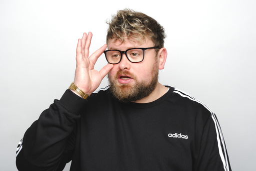 Photo of comedian William Stone; he adjusts his glasses, and is wearing a black Adidas jumper and a gold watch.