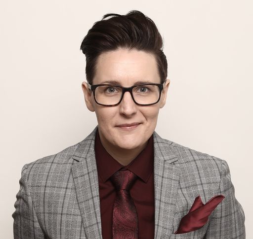 Portrait photograph of Sian Davies, wearing a grey checked suit, burgundy shirt and black glasses