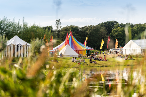 The festival site at Jupiter Rising. Two large tents, one with blue stripes and one in a rainbow pattern, are visible through long grass. A lake sits to their right, with flags behind.